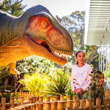 Two cute kids stand in the East Garden at Cal Academy while a life-size animatronic T. rex looms in the background