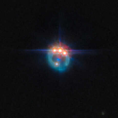 A small image of a galaxy distorted by gravitational lensing into a dim ring. At the top of the ring are three very bright spots with diffraction spikes coming off them, right next to each other: these are copies of a single quasar in the lensed galaxy, duplicated by the gravitational lens. In the centre of the ring, the elliptical galaxy doing the lensing appears as a small blue dot. The background is black and empty.