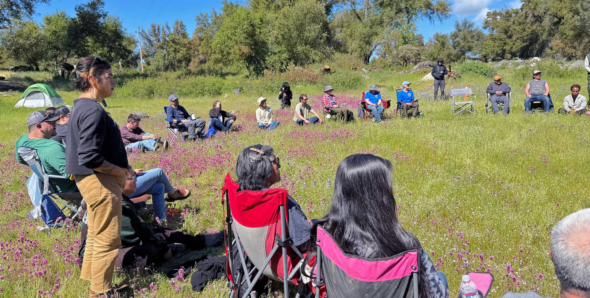 A group of 20 or so people sit and stand in a circle on a grassy, flowery meadow, during the fire ceremony.
